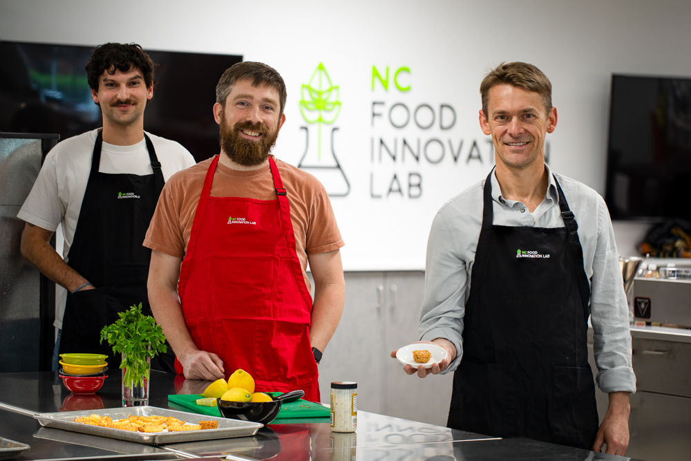 Three men stand in the NCFIL test kitchen with a plate of fish nuggets from the Atlantic Fish Co.